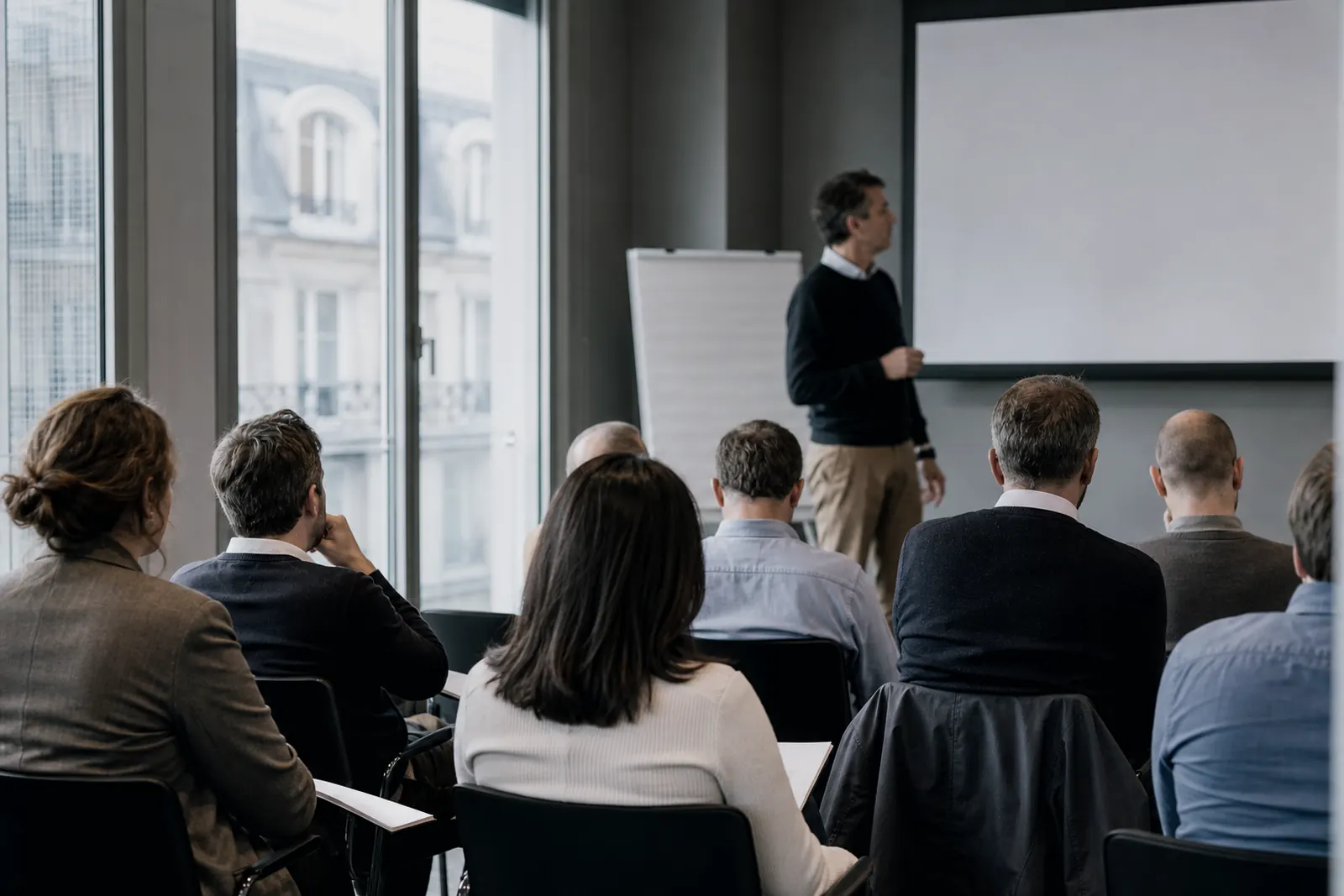 Salle de formation professionnelle : des adultes assis suivent un intervenant debout près d’un tableau.